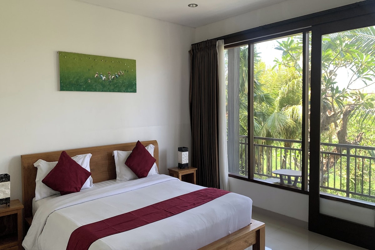 Quarto de hotel com uma cama grande, uma mesinha de cabeceira, um quadro na parede e uma janela com vista para o verde.
