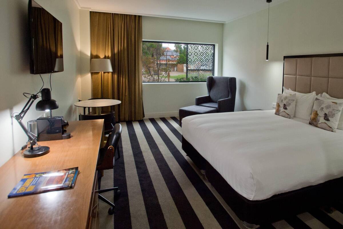 A hotel room with a bed, a desk with a lamp, a chair, a window with curtains, and a view of the outside.