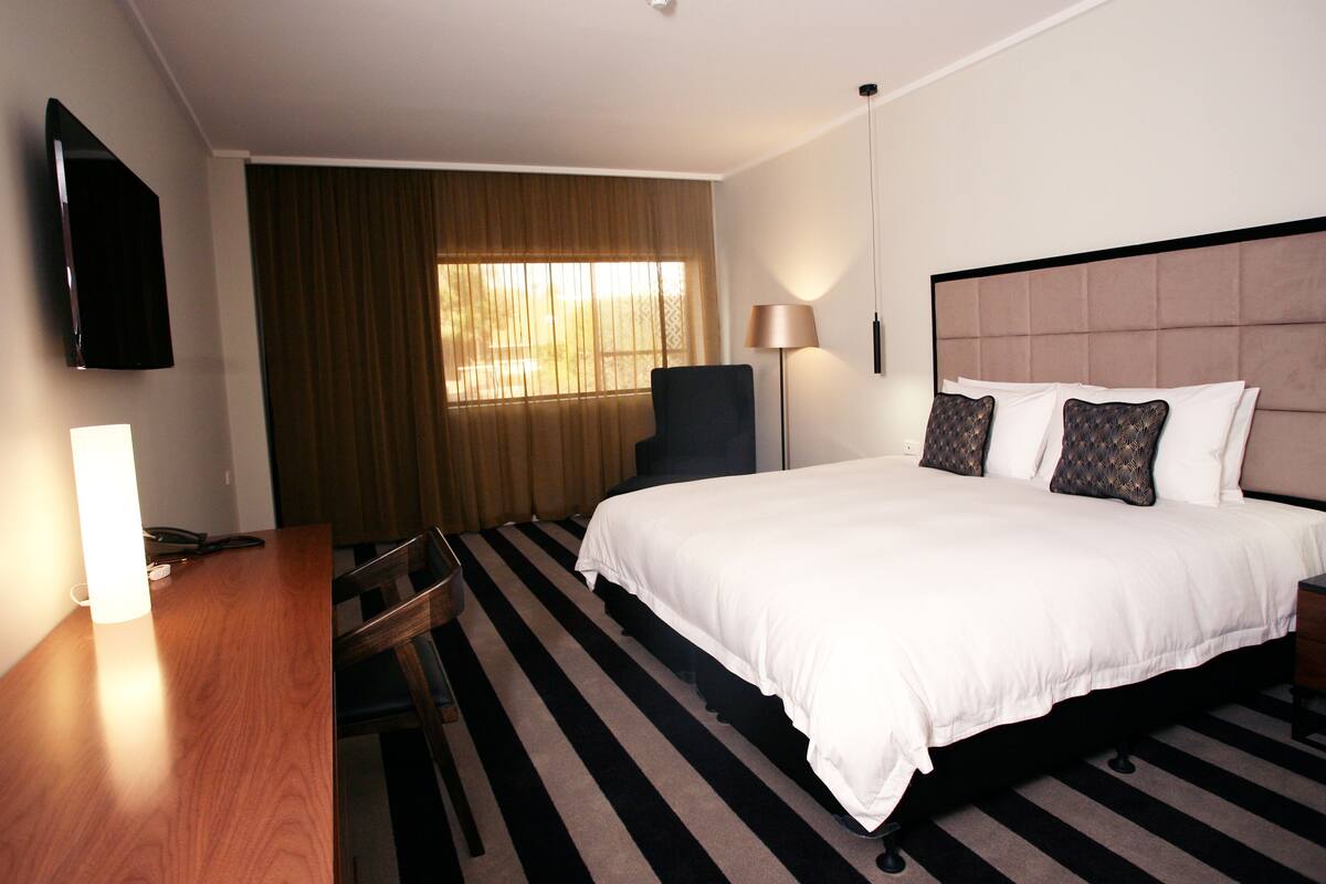 A hotel room with a bed, a desk, a chair, a TV, a lamp, and a window with curtains.