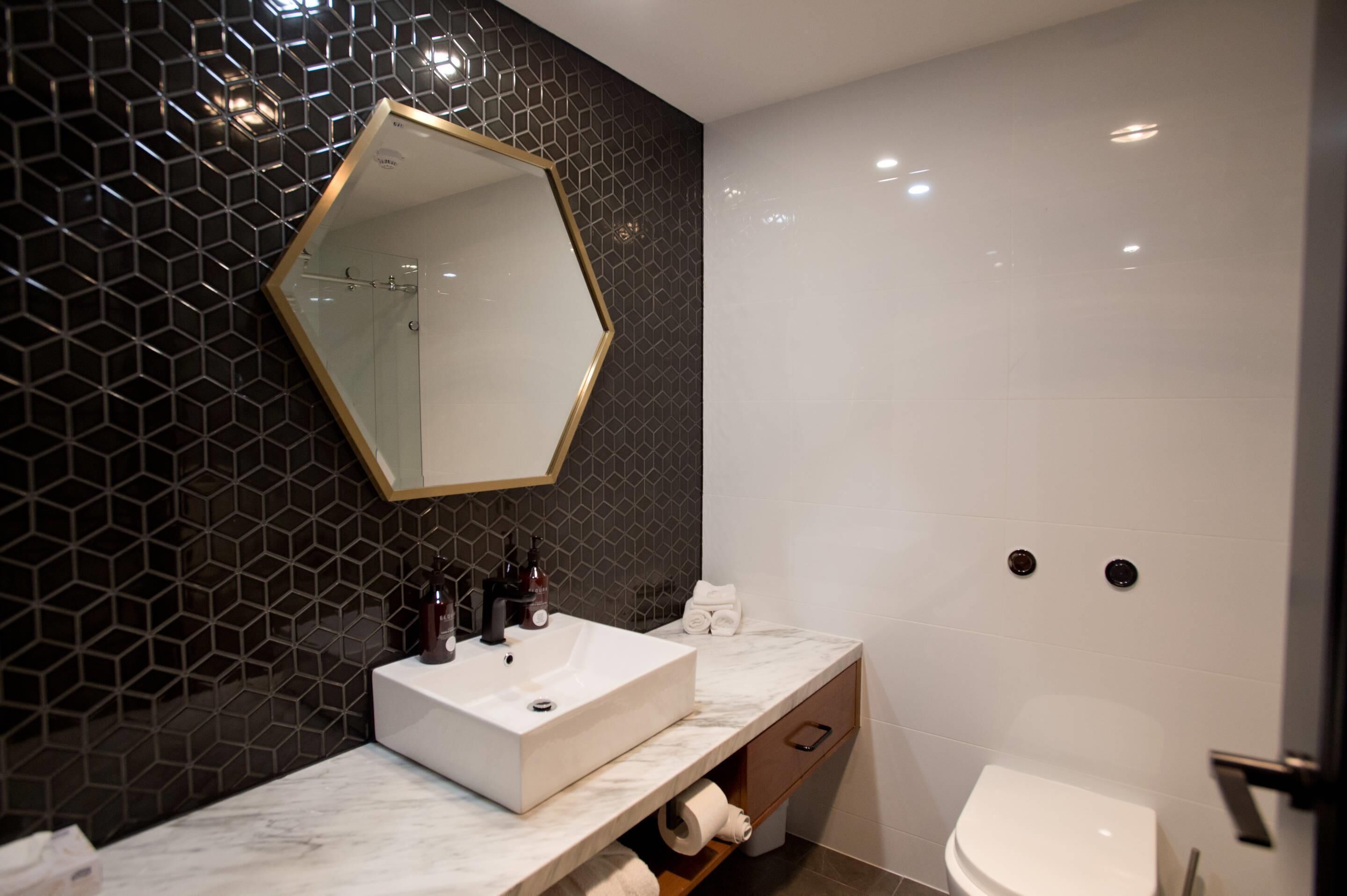 A modern bathroom with a hexagonal mirror, a white sink, and a toilet.