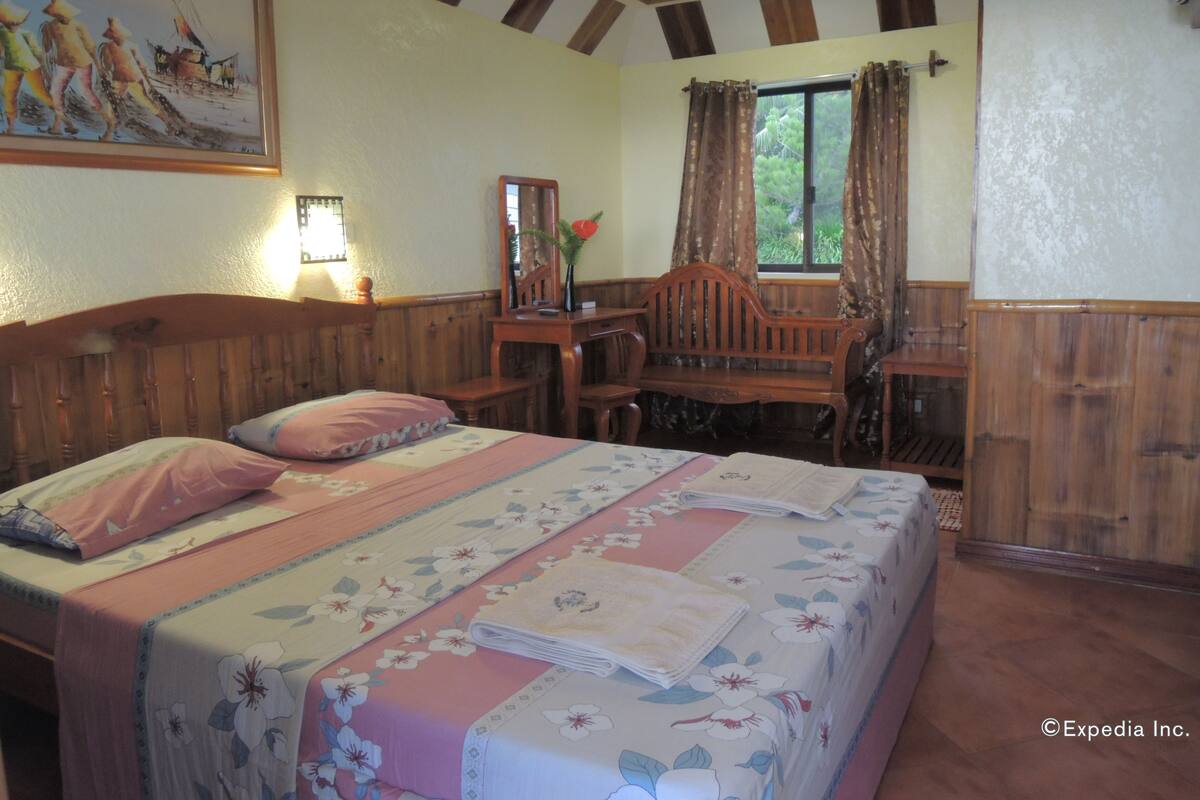 A bedroom with a wooden bed, a desk, and a chair. There is a window with curtains, a painting on the wall, and a lamp on the bedside table.