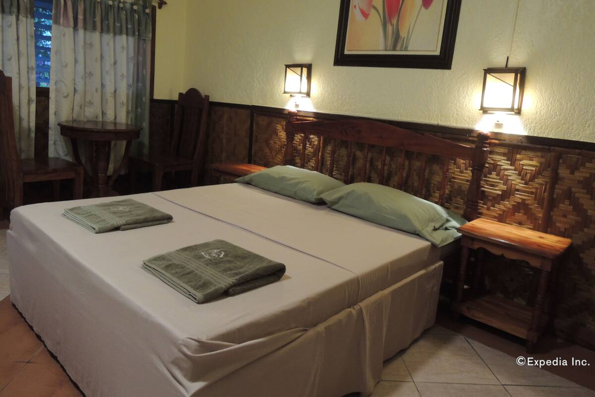 A double bed with two pillows, a wooden headboard, two bedside tables with lamps, a small wooden table, and a chair.