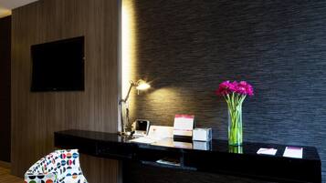 A reception desk with a flat-screen TV, a desk lamp, a vase with pink flowers, and a chair with a colorful pattern.