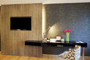 A modern reception area with a flat-screen TV mounted on a wooden panel wall, a desk with a lamp, a vase with flowers, and a patterned chair.