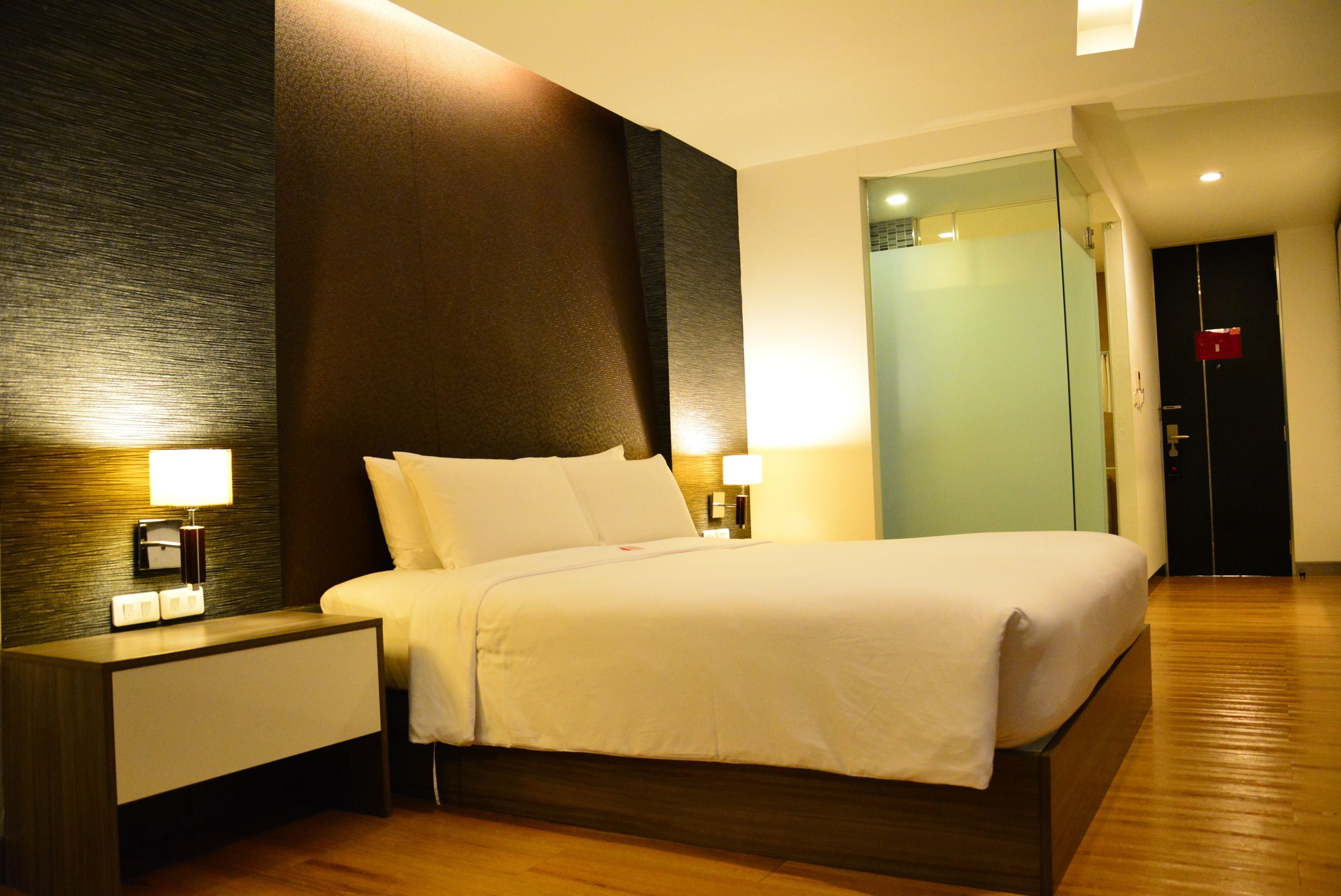 A hotel room with a large bed, two bedside tables with lamps, a wooden floor, and a glass door leading to a bathroom.