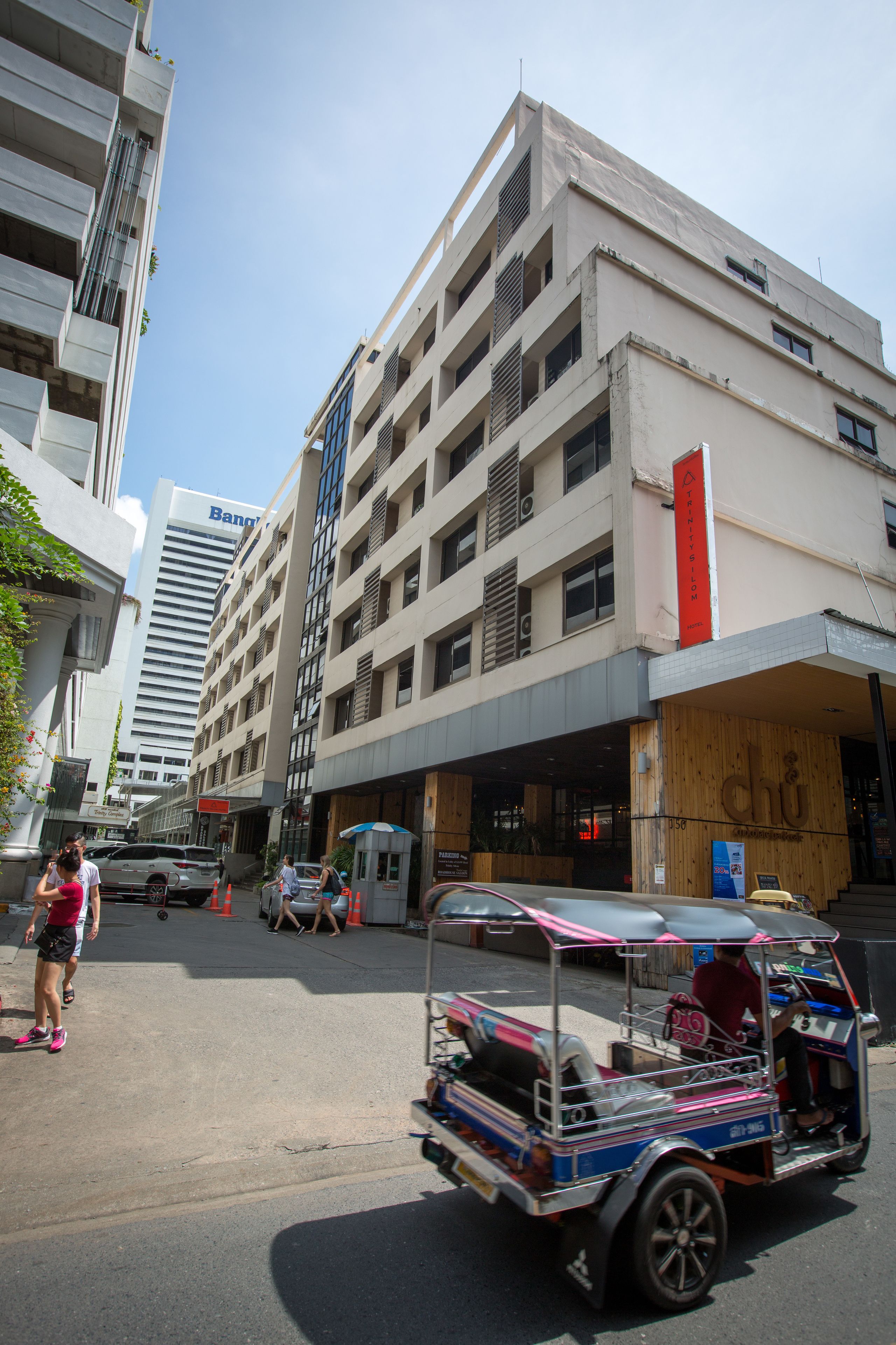 A multi-story building with a prominent red sign, a tuk-tuk on the street, and a clear blue sky.