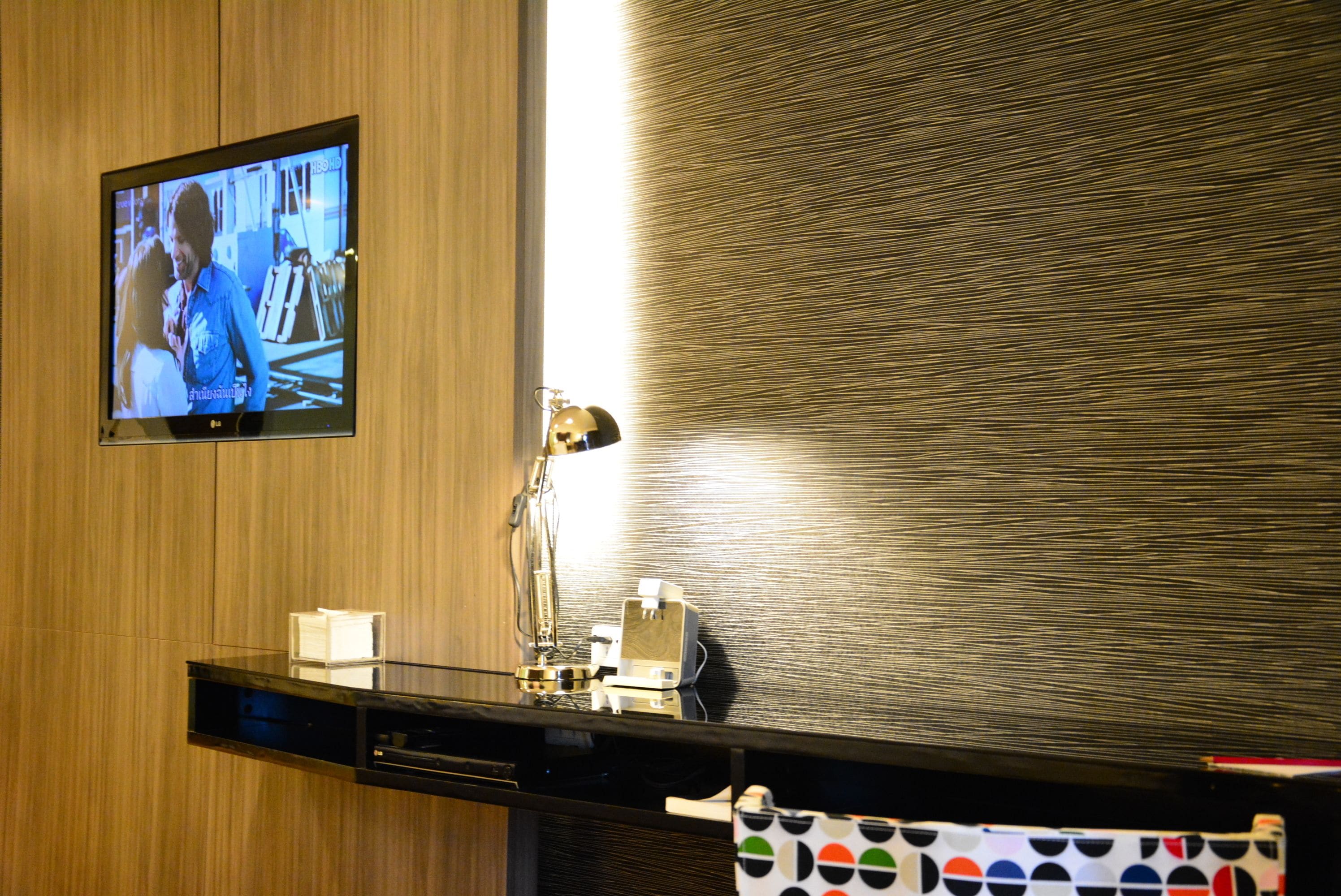 A hotel room with a wooden wall, a flat-screen TV mounted above a black desk, a desk lamp, and a towel with a polka dot pattern.