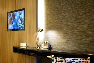 Hotelkamer met een houten wand, een flatscreen televisie boven een zwart bureau, een bureaulamp en een handdoek met stippen.