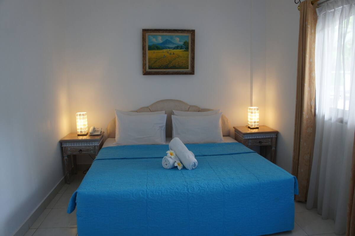 A neatly made bed with a blue bedspread, two bedside lamps, a painting on the wall, and a window with curtains.