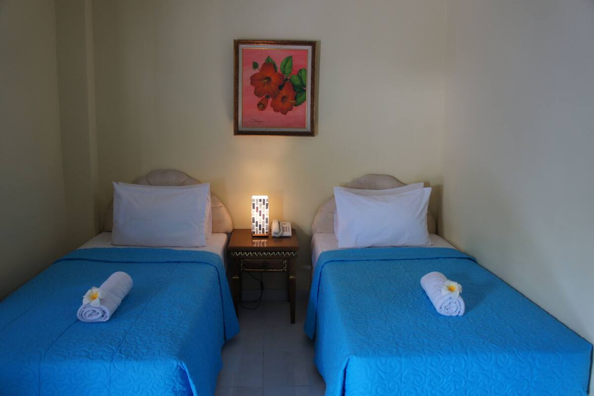 A hotel room with two single beds, a nightstand with a lamp, and a framed picture on the wall.