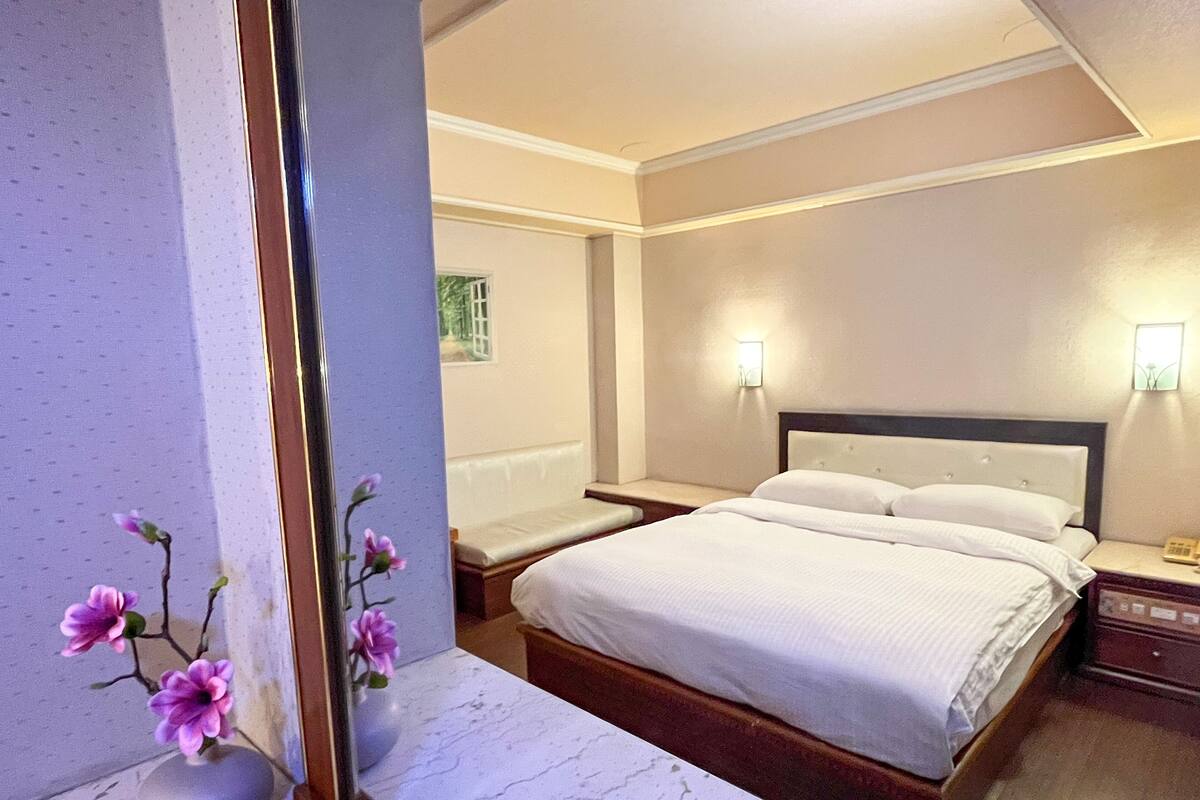 Habitación de hotel con una cama grande, mesitas de noche y un florero con flores sobre una encimera de mármol.