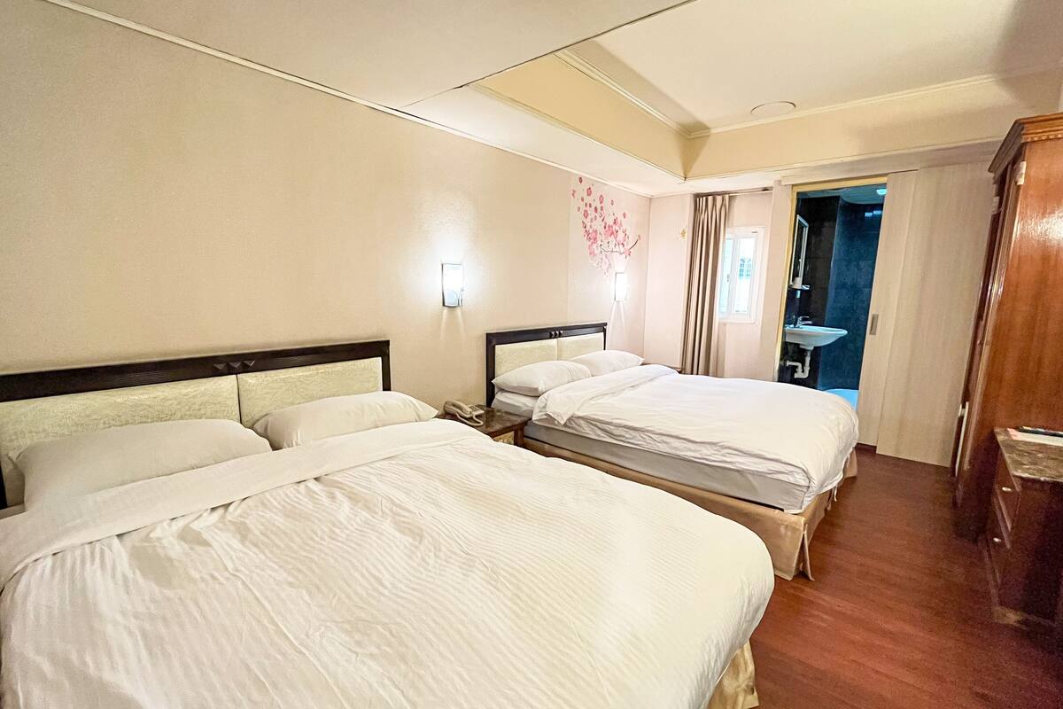 Habitación de hotel con dos camas, una mesita de noche con lámpara, baño con lavamanos y una puerta que da a otra habitación.