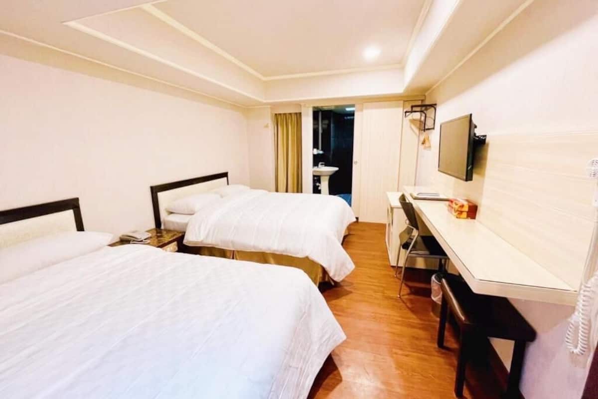 Habitación de hotel con dos camas, un escritorio, una silla, un televisor y un baño visible a través de una puerta abierta.