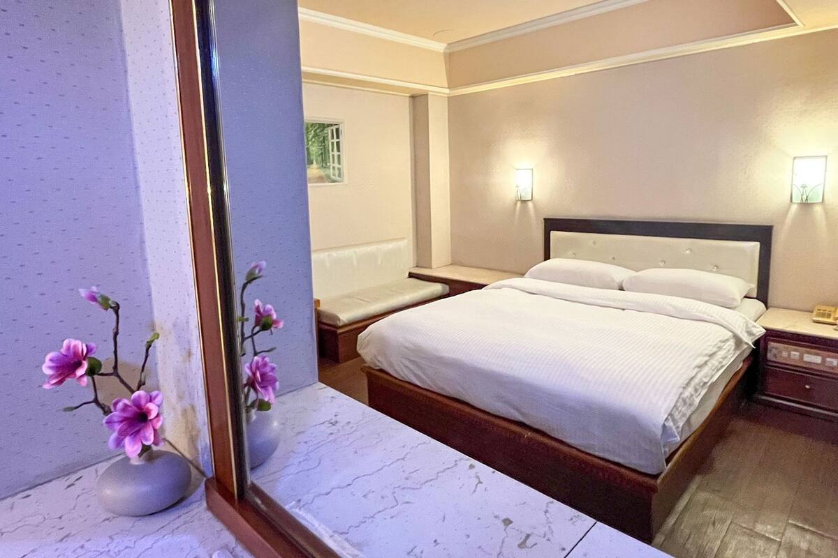 Una habitación de hotel con una cama grande, mesitas de noche, un espejo y un jarrón con flores.