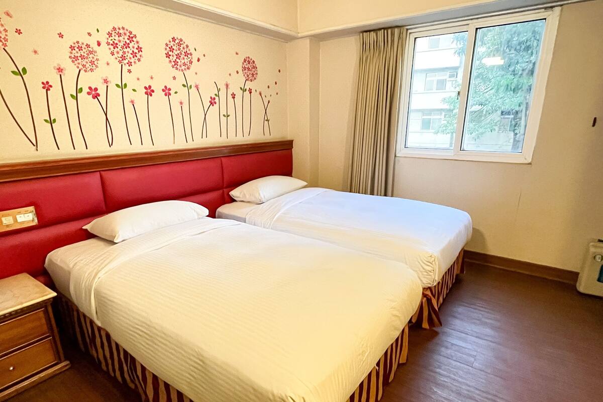 Habitación de hotel con dos camas, cabecero rojo, piso de madera y una ventana con cortinas.