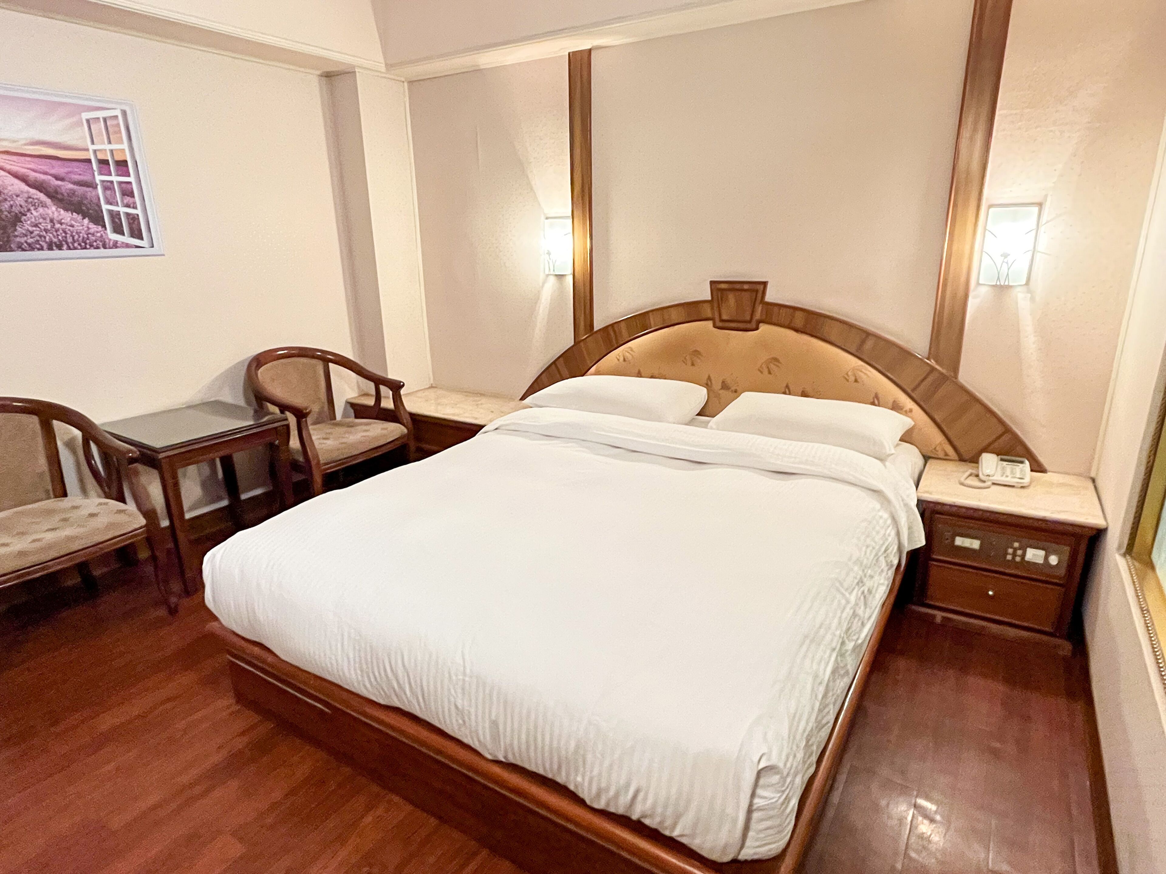 A hotel room with a large bed, a wooden nightstand, a desk with a chair, and a wall-mounted lamp.