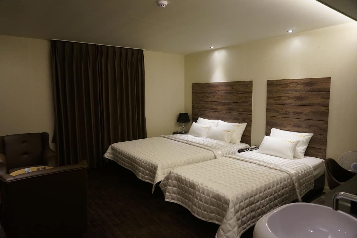 A hotel room with two beds, a desk, a chair, a large window with curtains, and a bathroom with a sink and mirror.