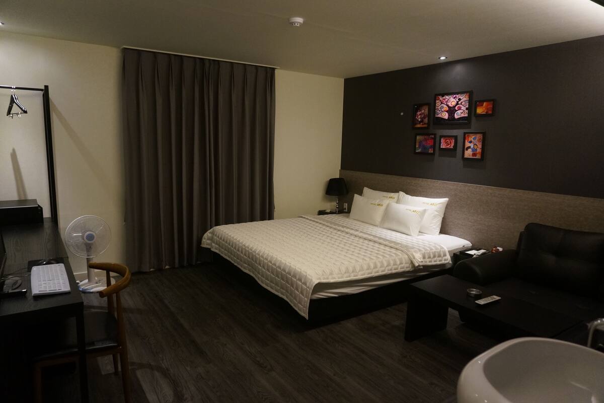 A hotel room with a bed, a desk, a chair, a fan, a couch, and artwork on the wall.