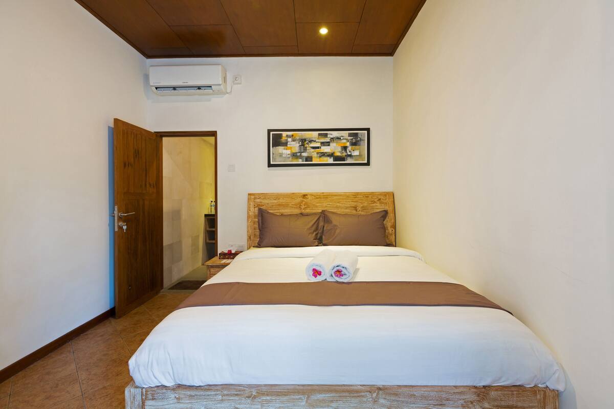 A hotel room with a large bed, a wooden headboard, a wall-mounted air conditioner, and a framed artwork above the bed.