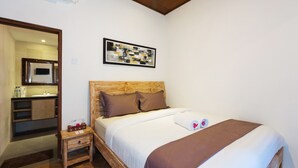 Habitación de hotel con cabecera de madera, una cama grande con ropa de cama blanca y marrón, una mesita de noche pequeña con un reloj, una obra de arte abstracta enmarcada sobre la cama, y un baño visible a través de una puerta abierta.