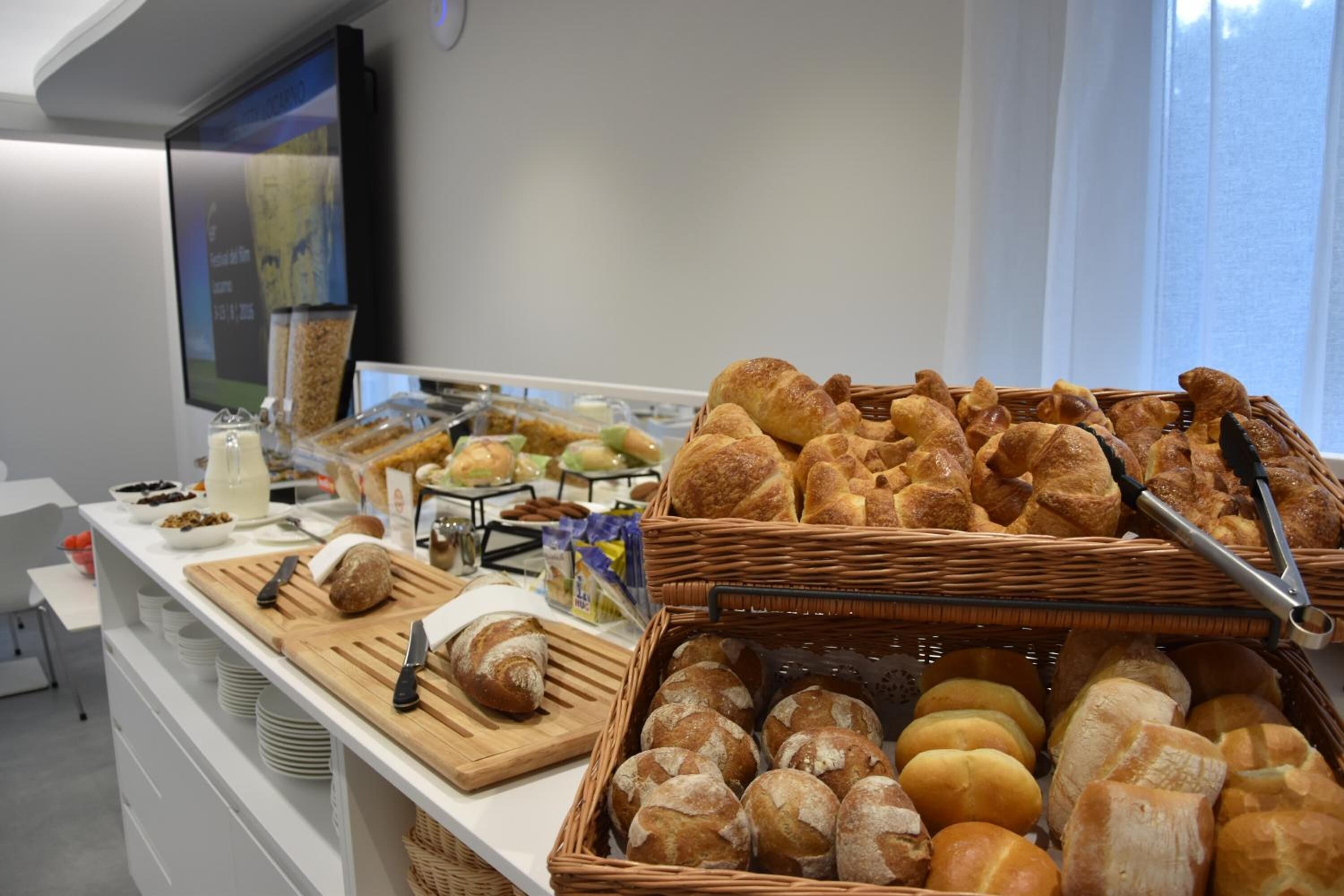 A buffet with various breads and pastries, a cutting board with a knife, and a television screen in the background.