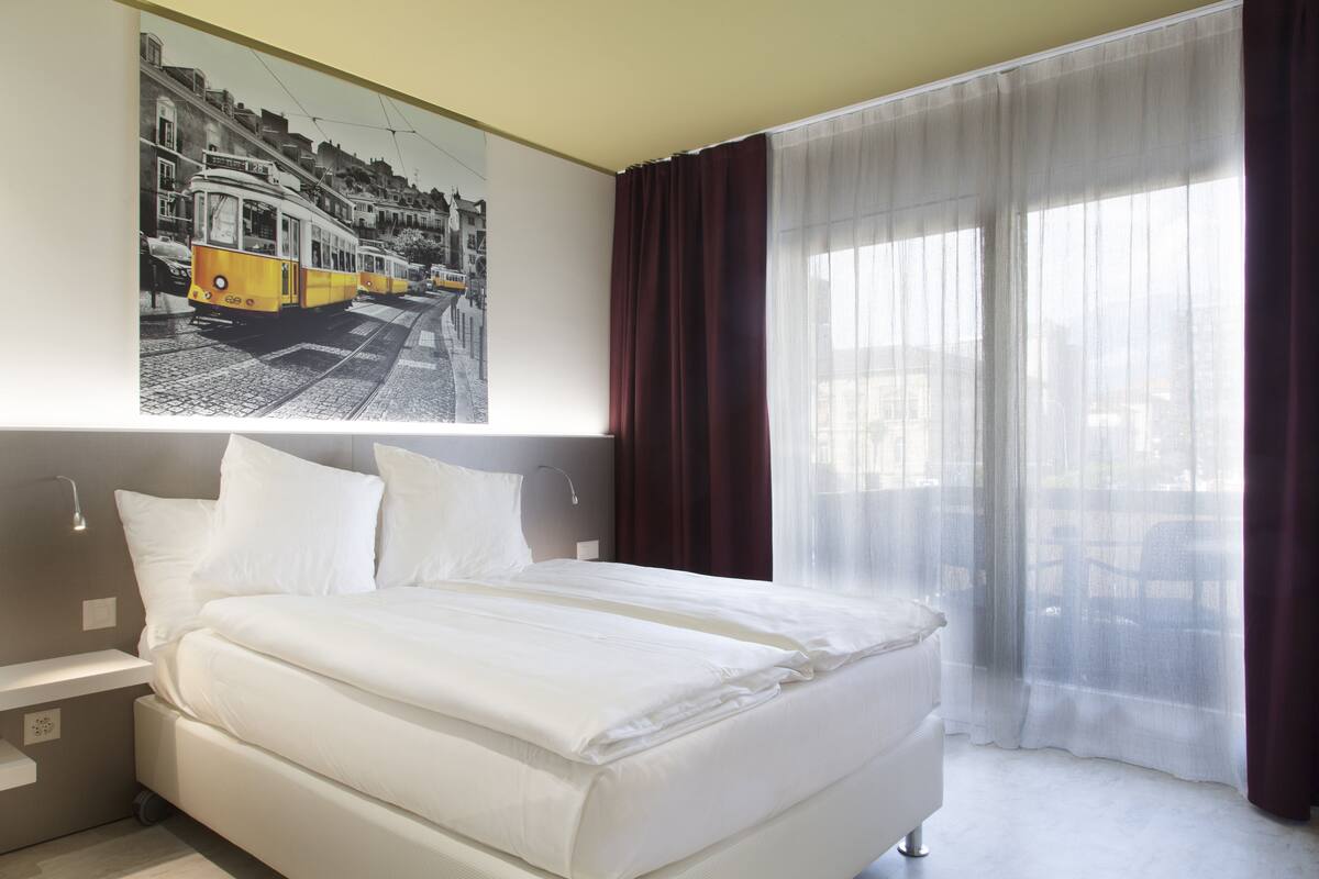 A modern hotel room with a large bed, a wall-mounted artwork, a window with sheer curtains, and a bedside table.