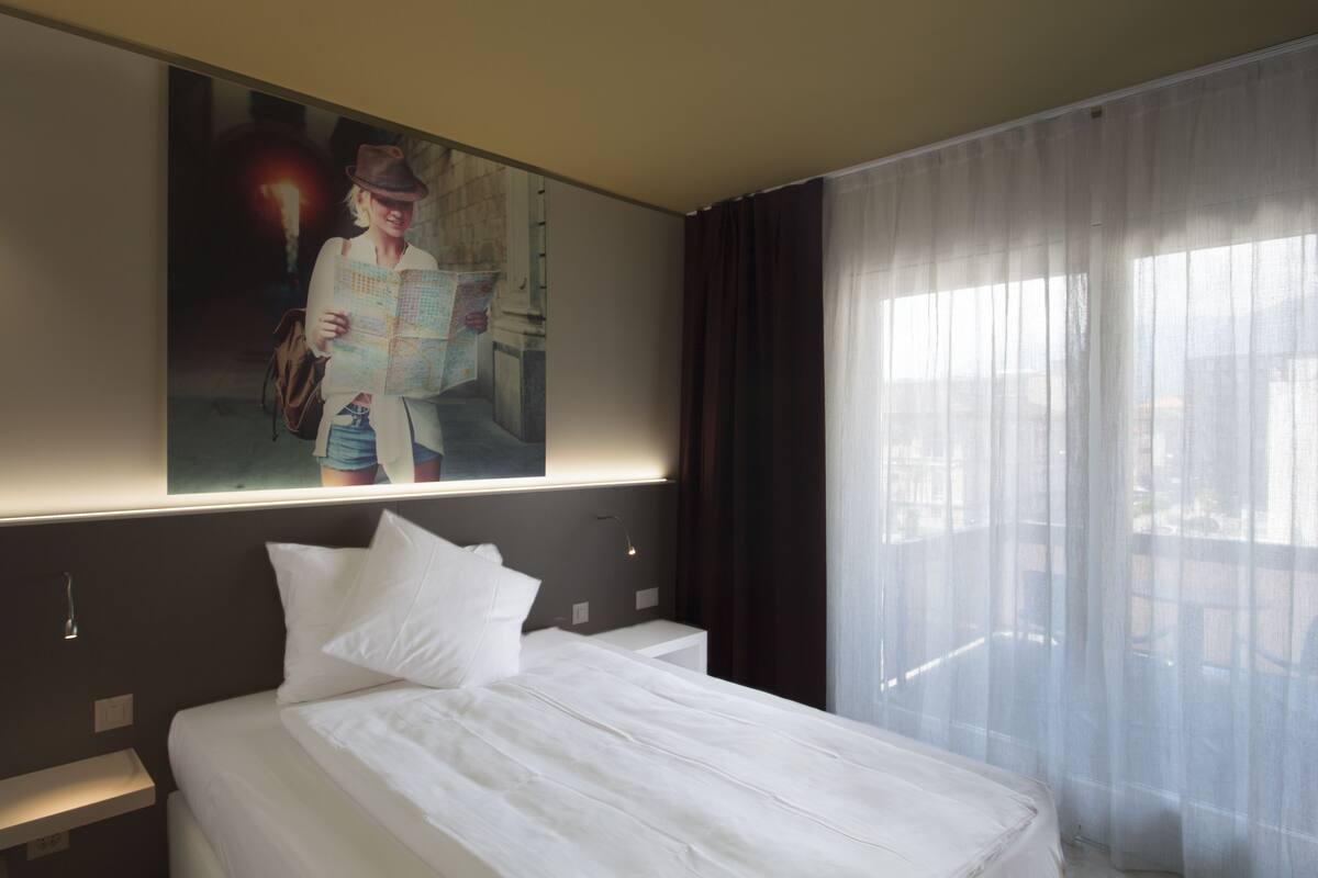 A hotel room with a large bed, a nightstand, a wall-mounted painting, and sheer curtains.