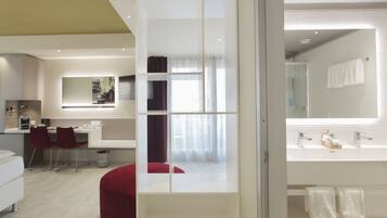 A modern hotel room with a desk, chairs, a red ottoman, a large mirror, and a bathroom with a sink and towel rack.