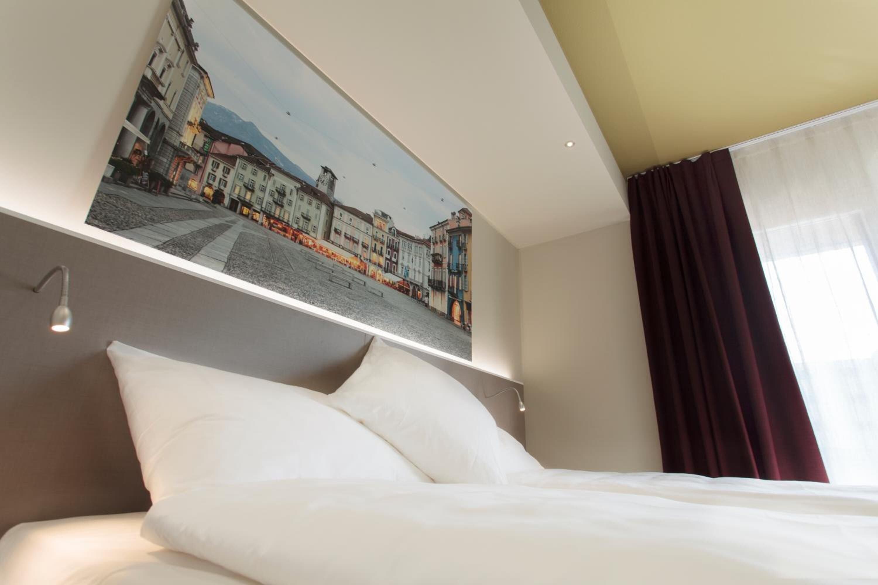 A hotel room with a bed, white pillows, a mounted picture of a street scene, and a window with curtains.