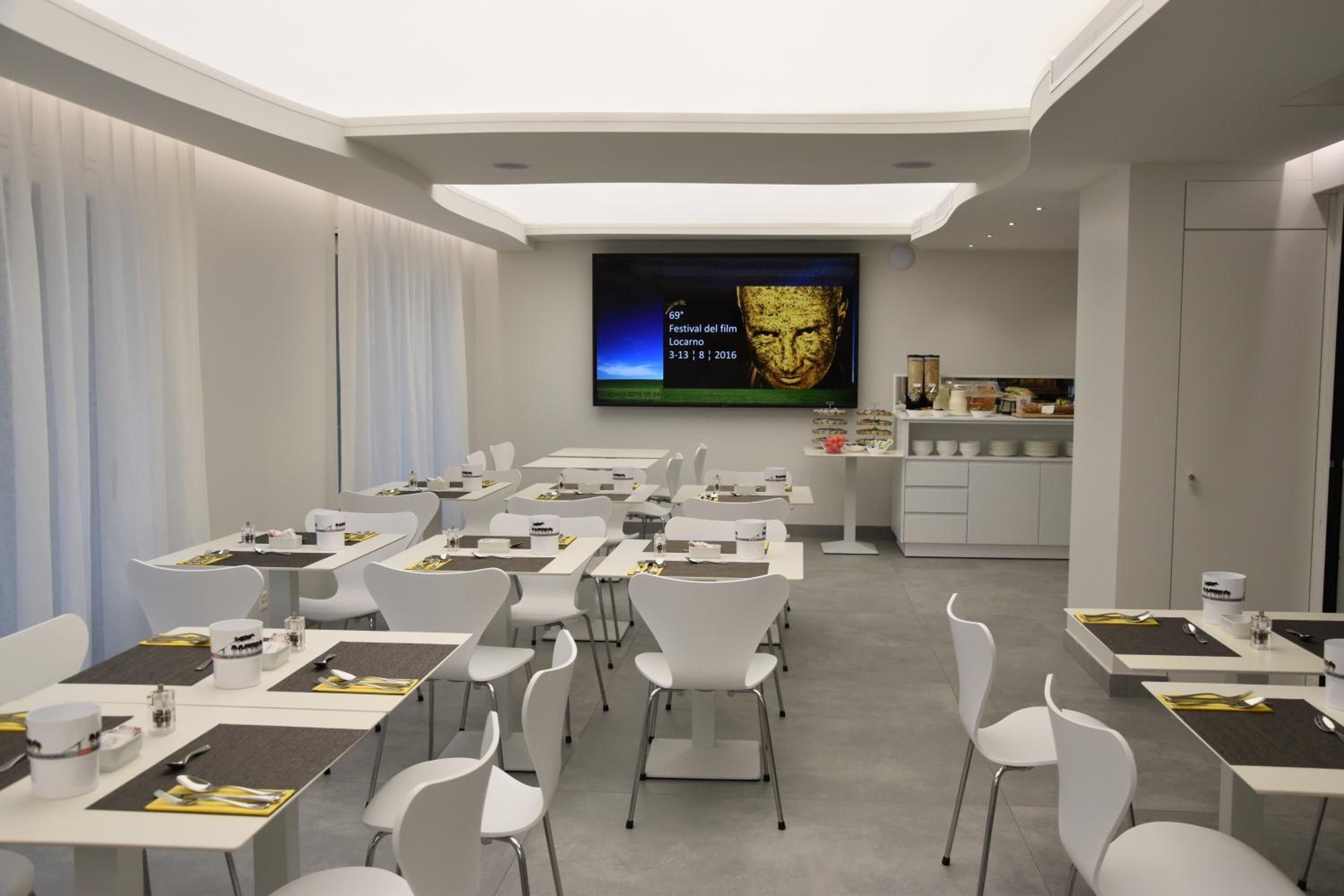 Una sala de conferencias moderna con mesas y sillas blancas, una pantalla grande que muestra un evento de festival de cine, y una zona de kitchenette.