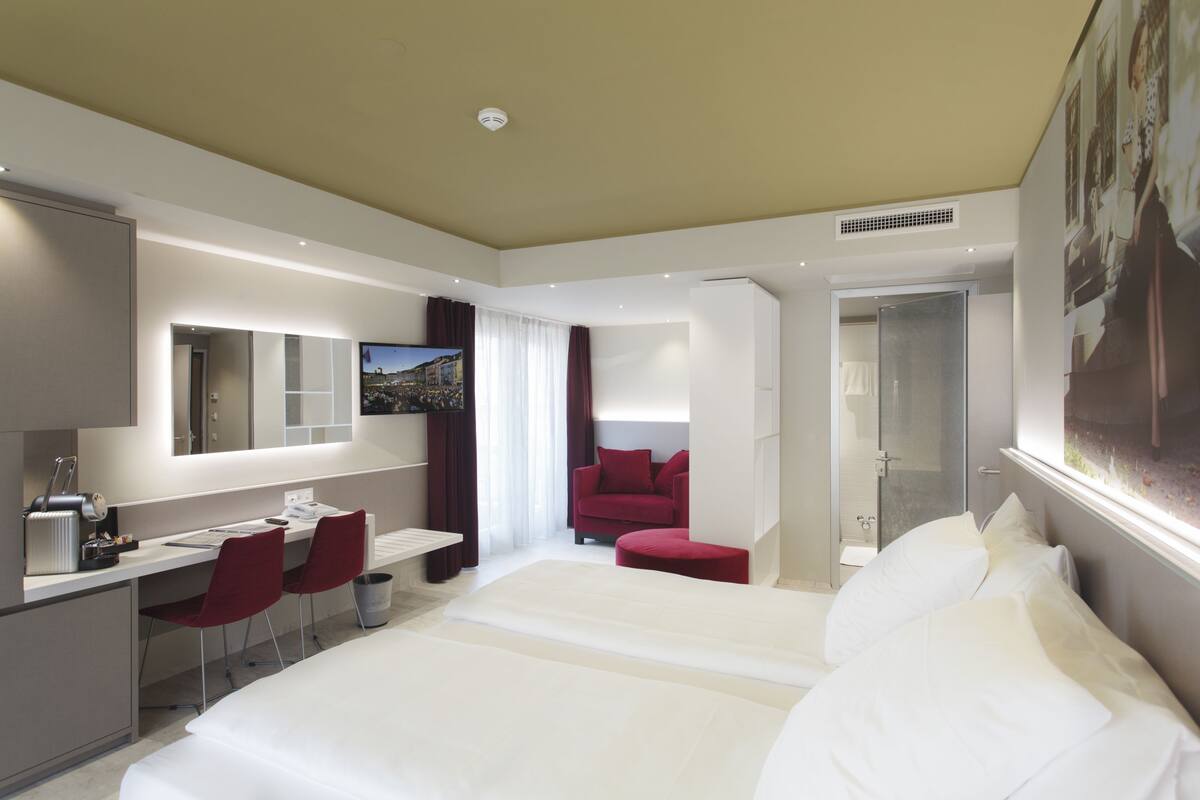 A modern hotel room with a large bed, a desk with two chairs, a TV, and a shower area.
