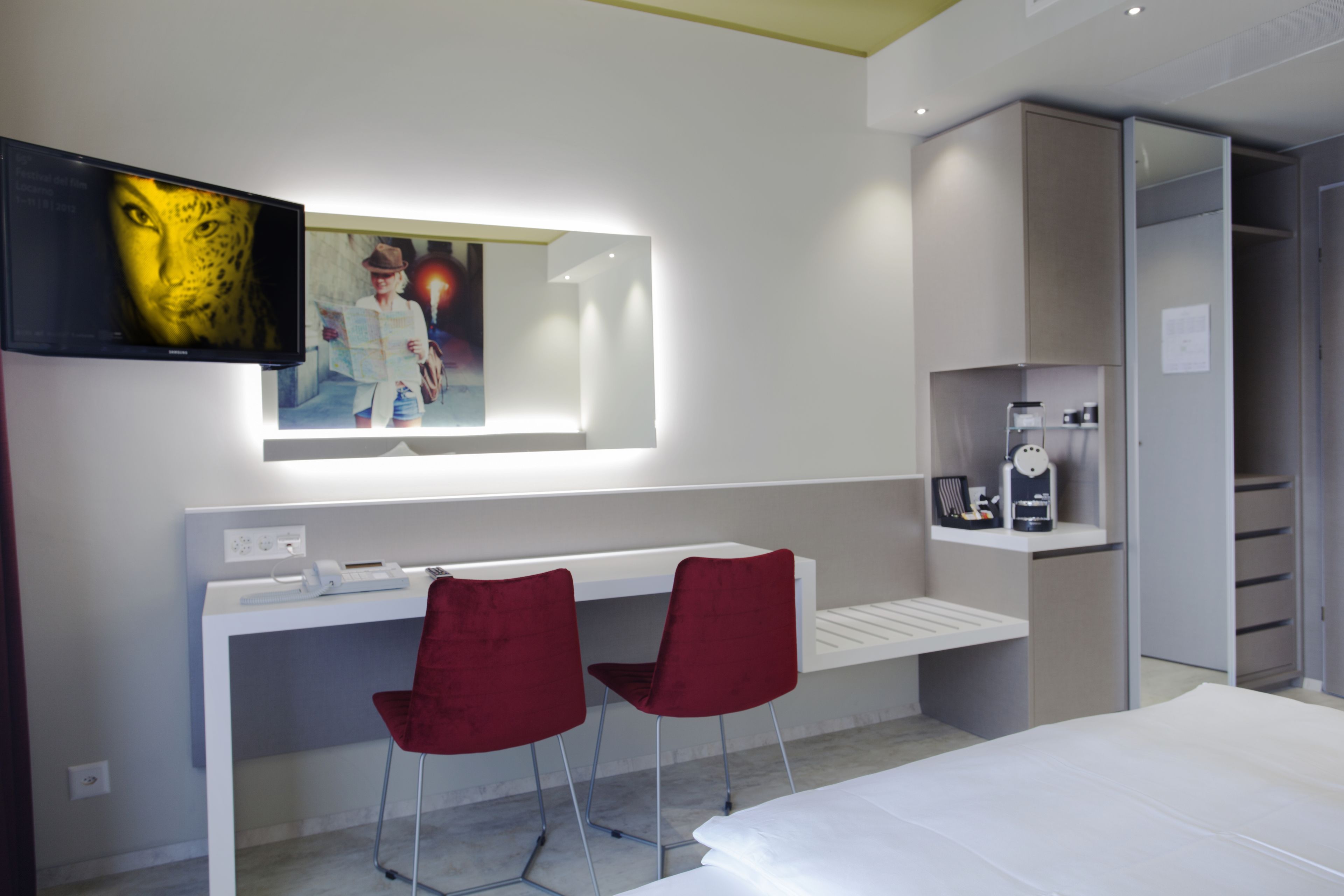 A modern hotel room with a white desk, two red chairs, a large mirror, and a bed.
