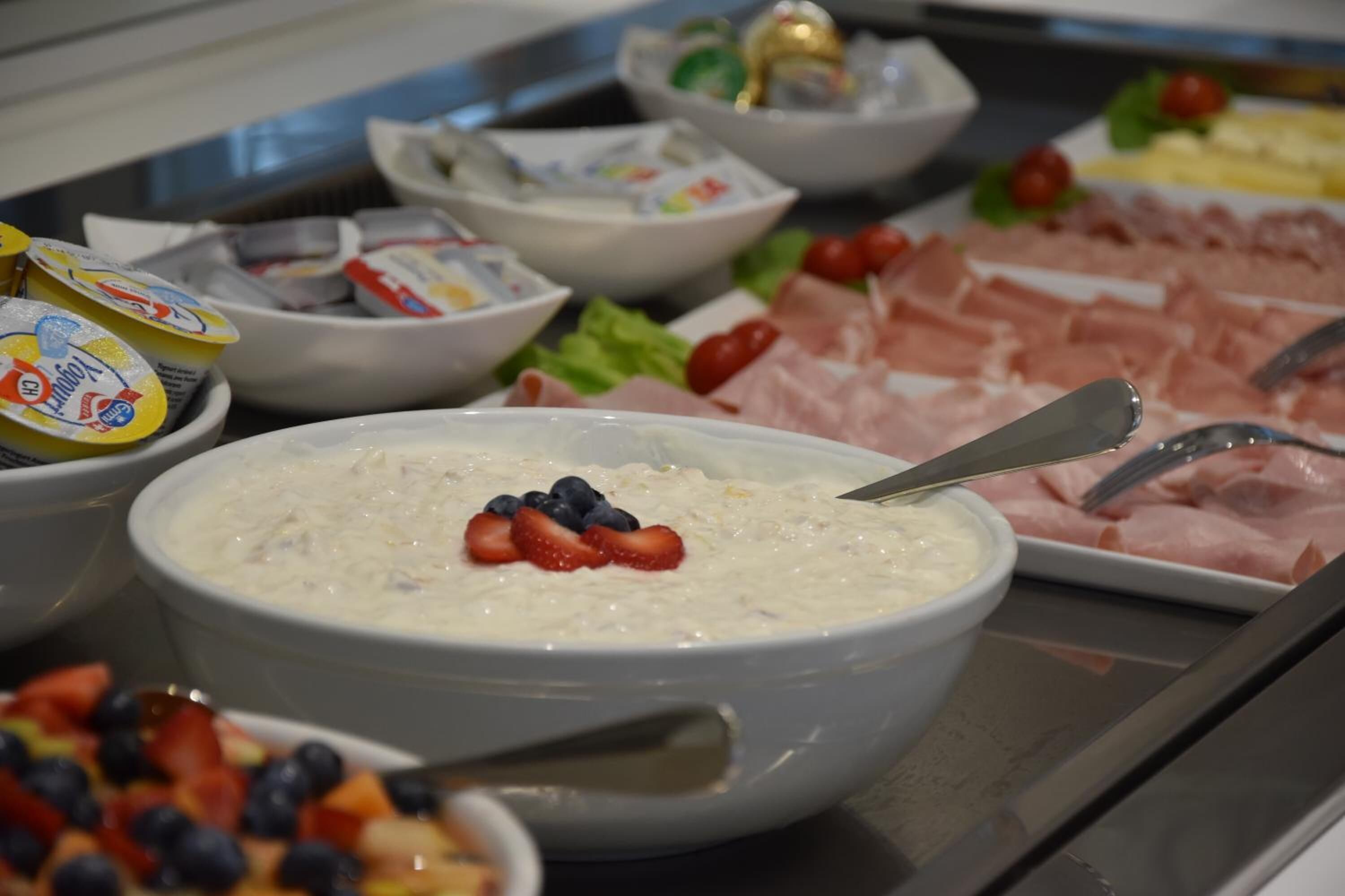 A buffet with various food items including a bowl of oatmeal with strawberries, bowls of yogurt, and a platter of sliced meats.
