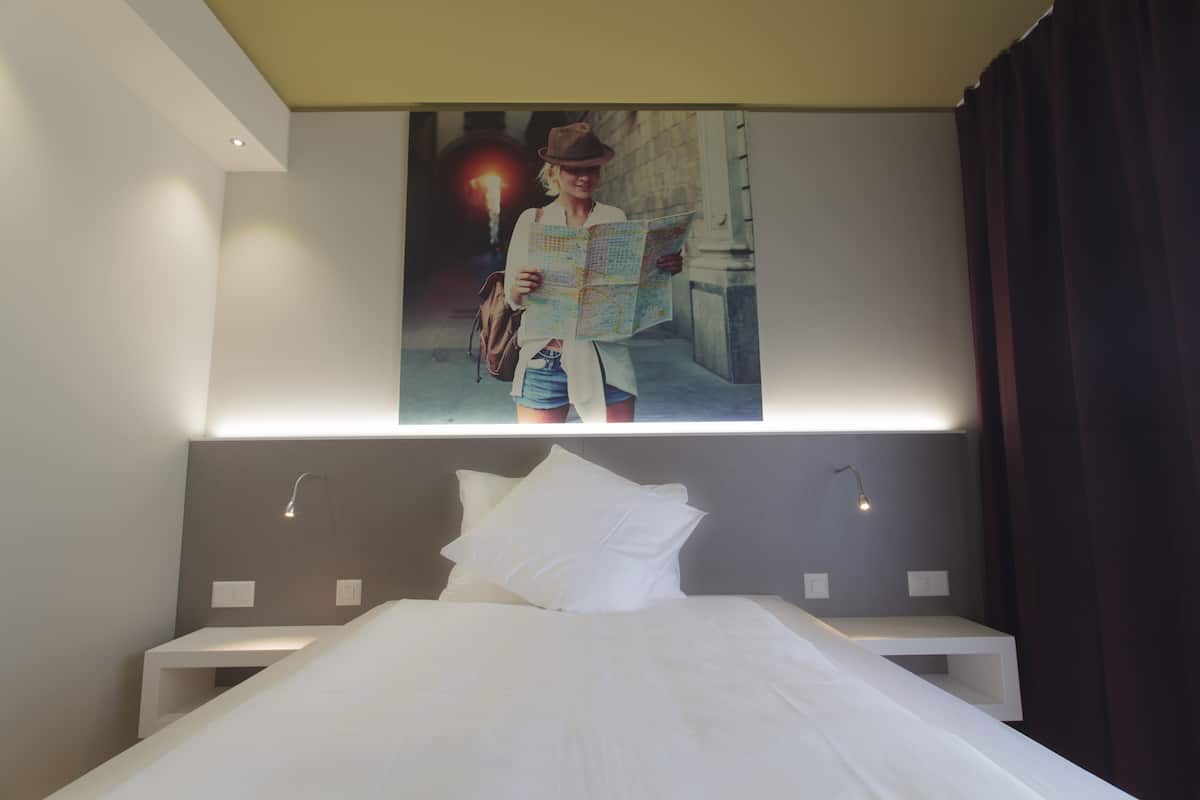 A hotel room with a large bed, a nightstand, a wall-mounted light, and a poster of a person holding a map.