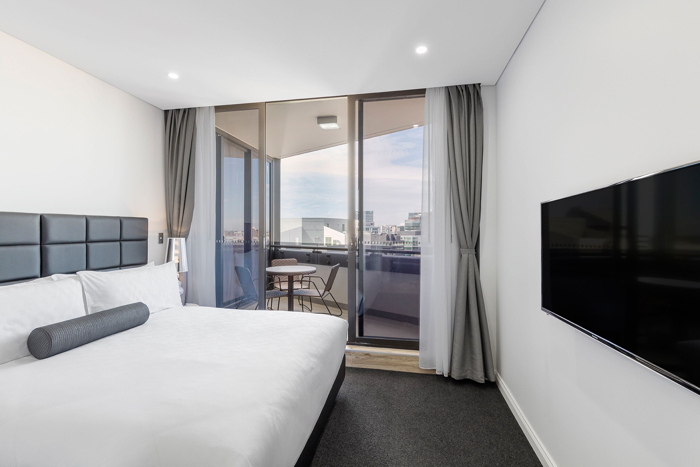 Habitación de hotel con una cama grande, un televisor de pantalla plana, un balcón con mesa y sillas, y vistas a la ciudad.