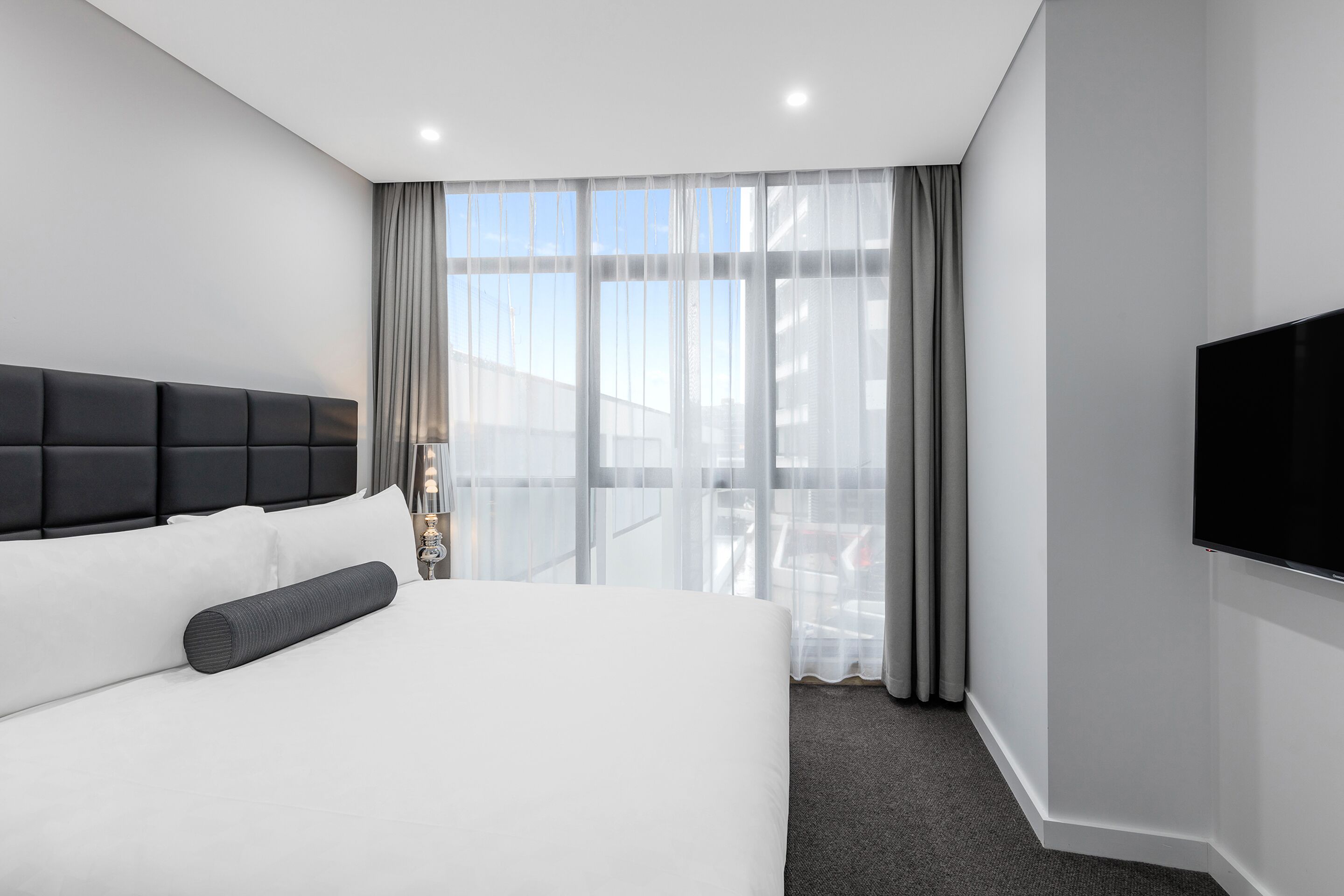 A modern hotel room with a large bed, a flat-screen TV mounted on the wall, and a view of the city through sheer curtains.