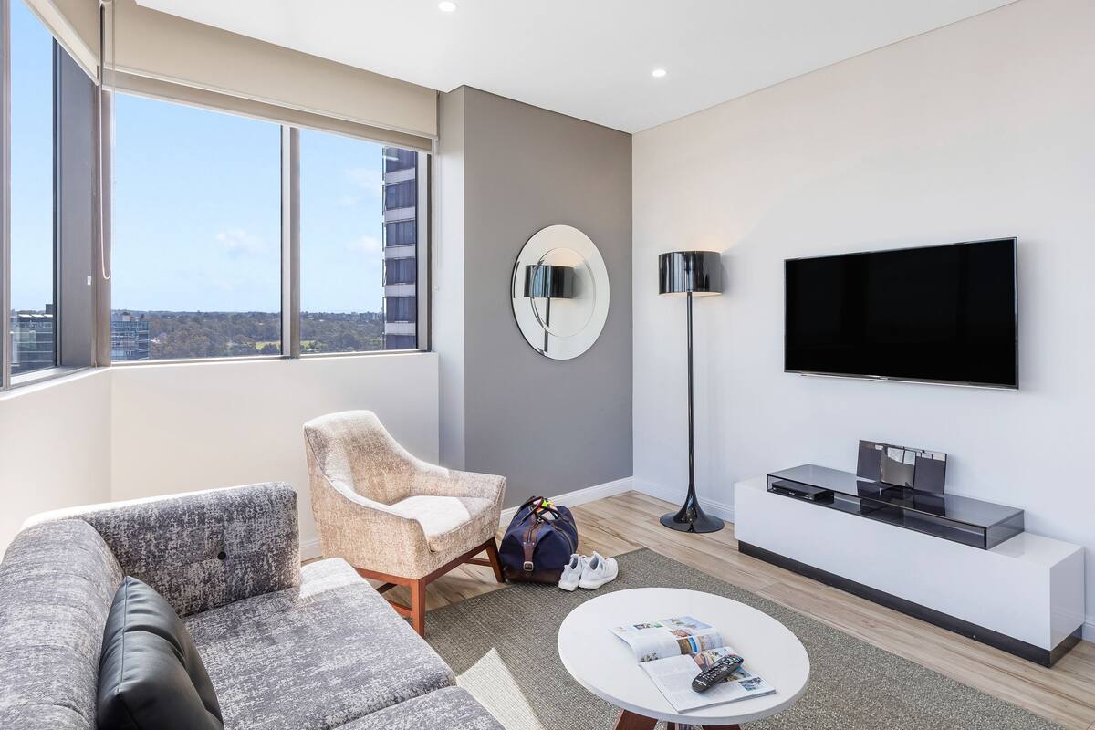 Ein modernes Wohnzimmer mit einer Couch, einem Sessel und einem Couchtisch, einem flachbildfernseher an der Wand und einem großen Fenster mit Blick auf die Stadt.