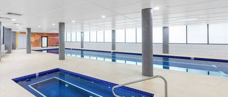 A swimming pool with a tiled deck, a metal handrail, and a view of the sky through large windows.