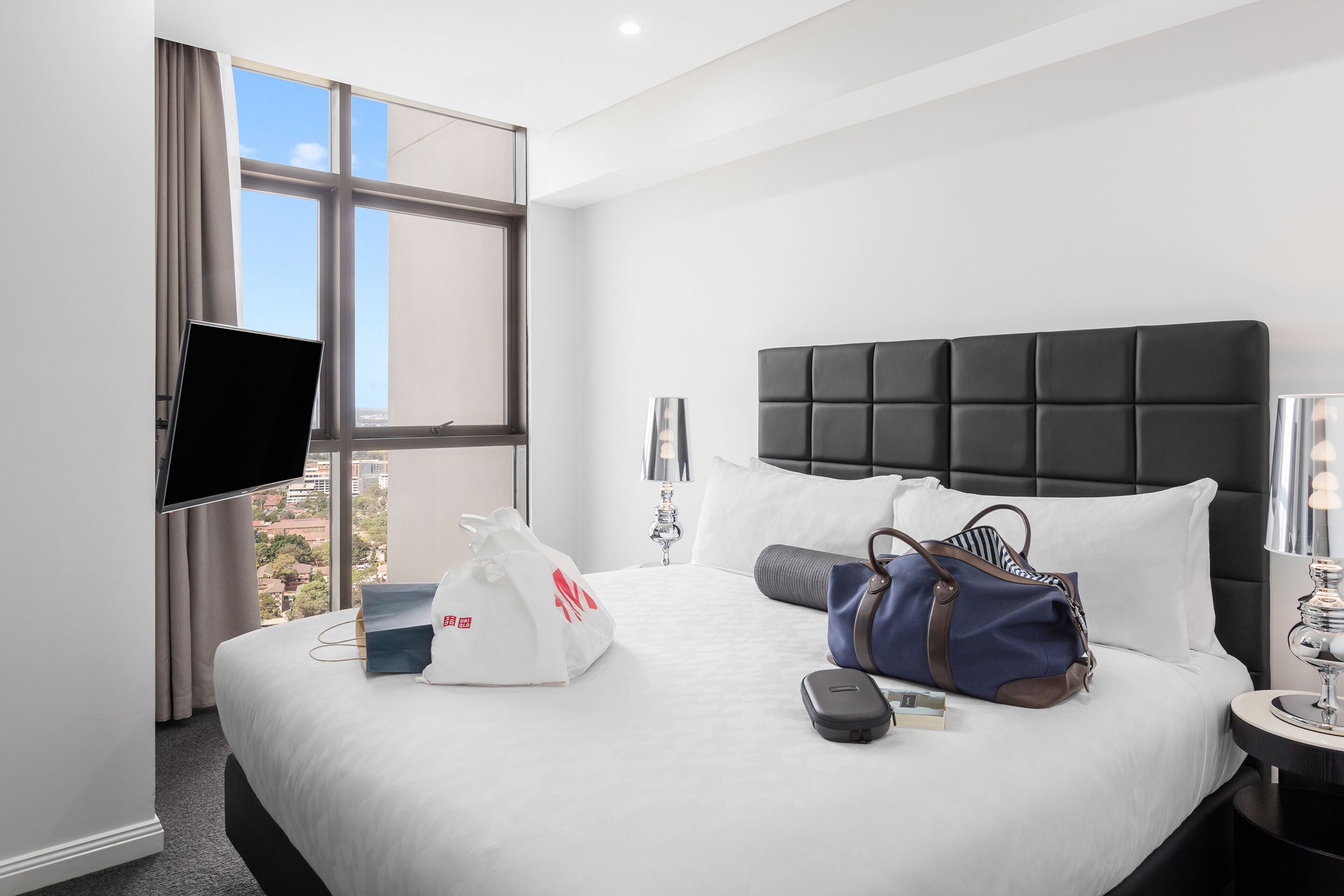 A hotel room with a large bed, a flat-screen TV mounted on the wall, a window with curtains, a bedside table with a lamp, and a view of the city.
