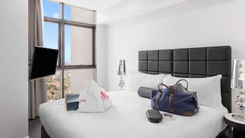 A hotel room with a large bed, a flat-screen TV mounted on the wall, a window with curtains, a bedside table with a lamp, and a view of the city.