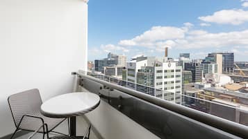 Un balcon avec une table blanche et des chaises en osier, donnant sur un paysage urbain avec des bâtiments modernes.