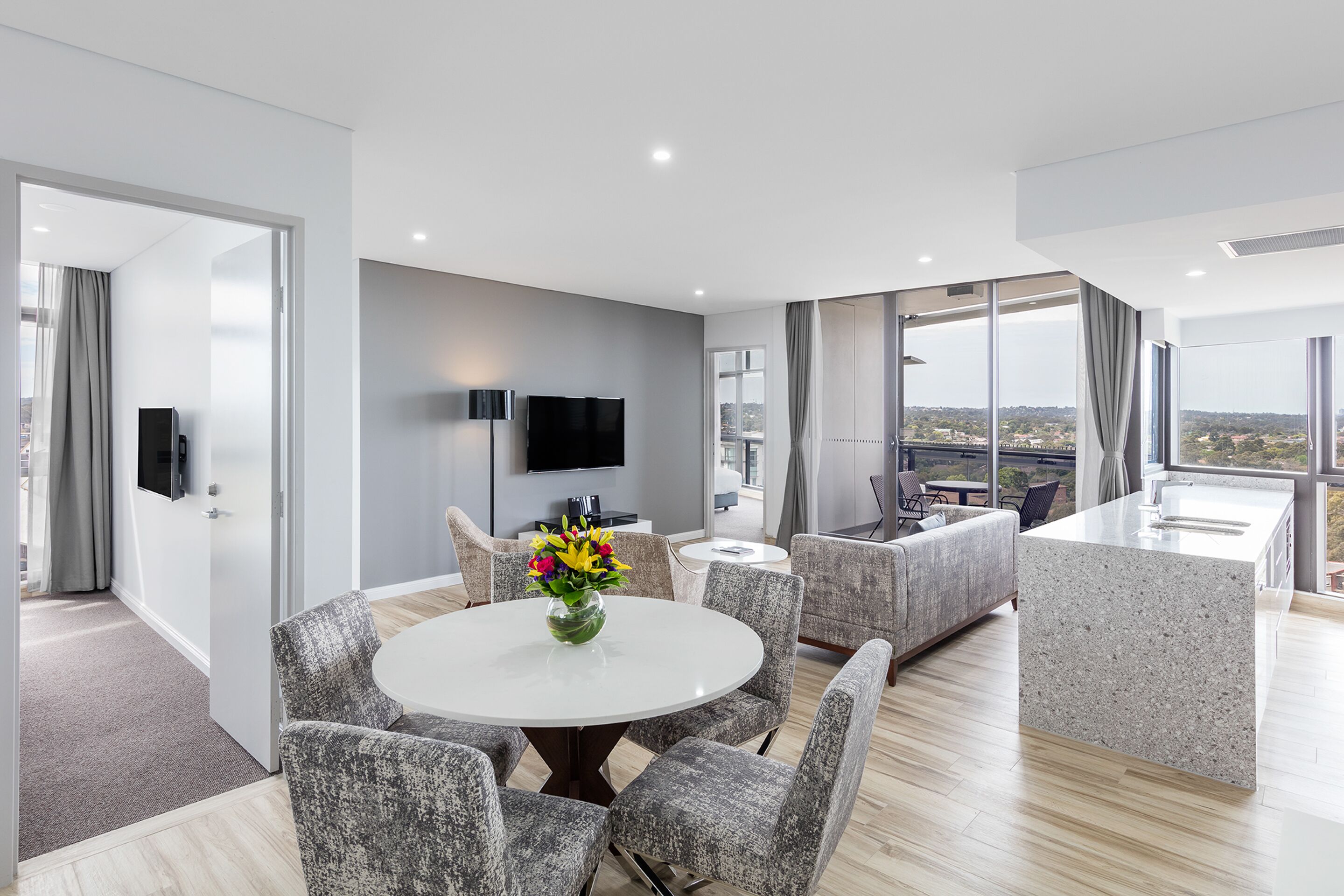 A modern living room with a round dining table, grey upholstered chairs, a flat-screen TV mounted on the wall, and a large window offering a city view.