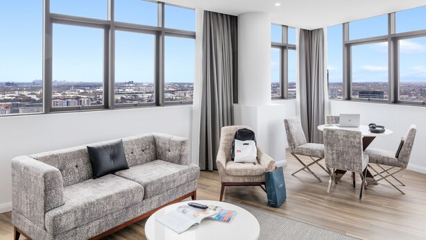 Una sala moderna con sofá, sillones, una mesa de centro y una mesa de comedor con una computadora portátil. Ventanas amplias ofrecen vista a la ciudad.