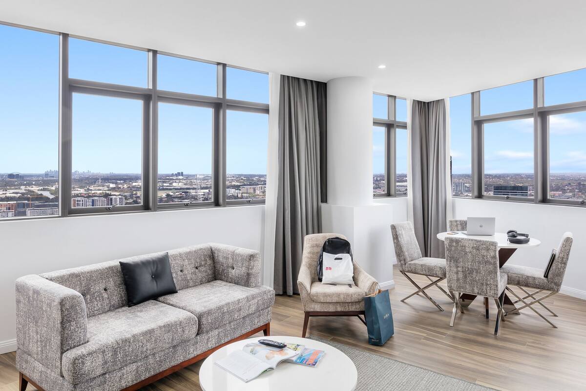 Ein modernes Wohnzimmer mit einer Couch, Sesseln, einem Couchtisch und einem Esstisch mit einem Laptop. Große Fenster bieten Blick auf die Stadt.