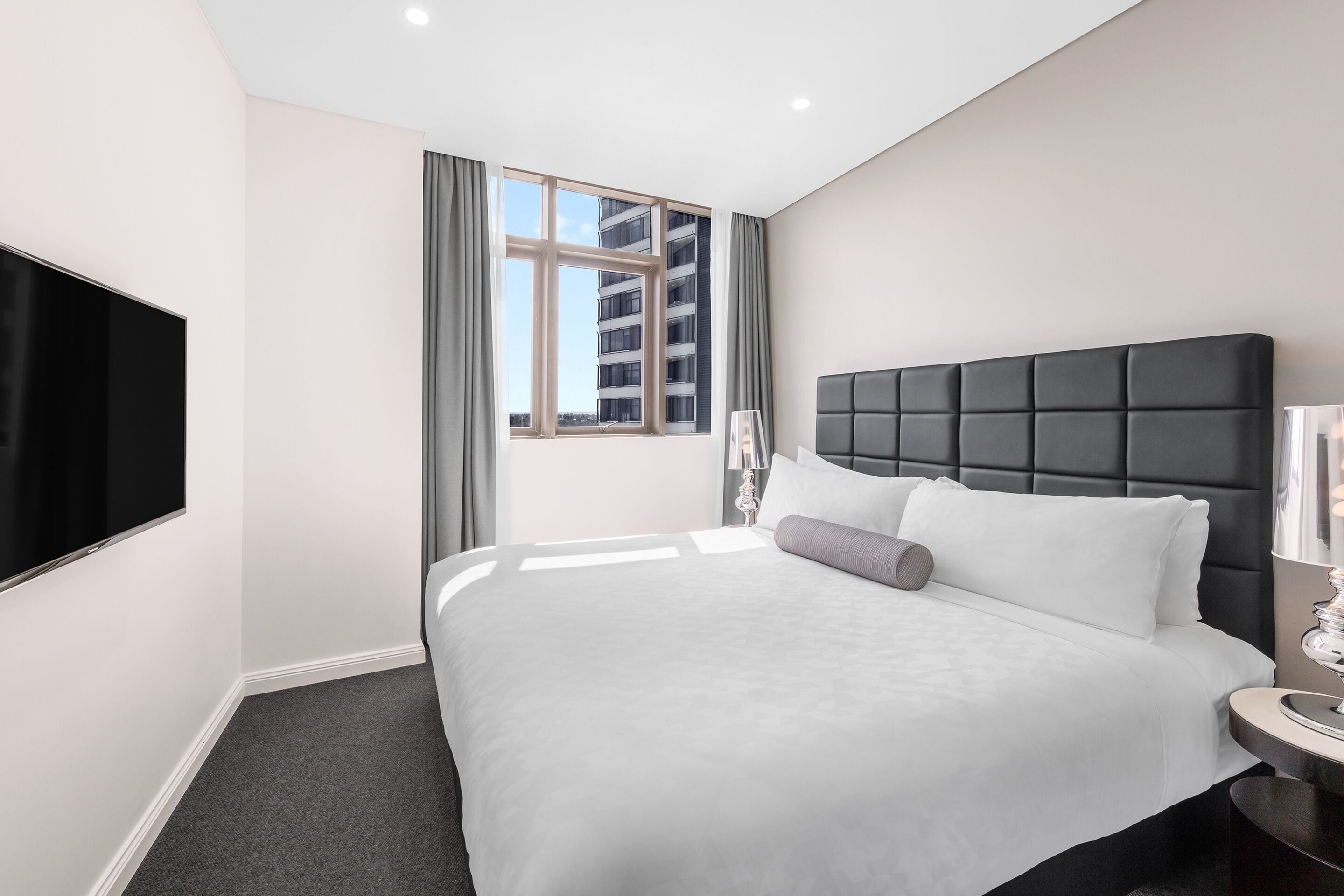 A modern hotel room with a large bed, a flat-screen TV, a window with curtains, and a nightstand with a lamp.