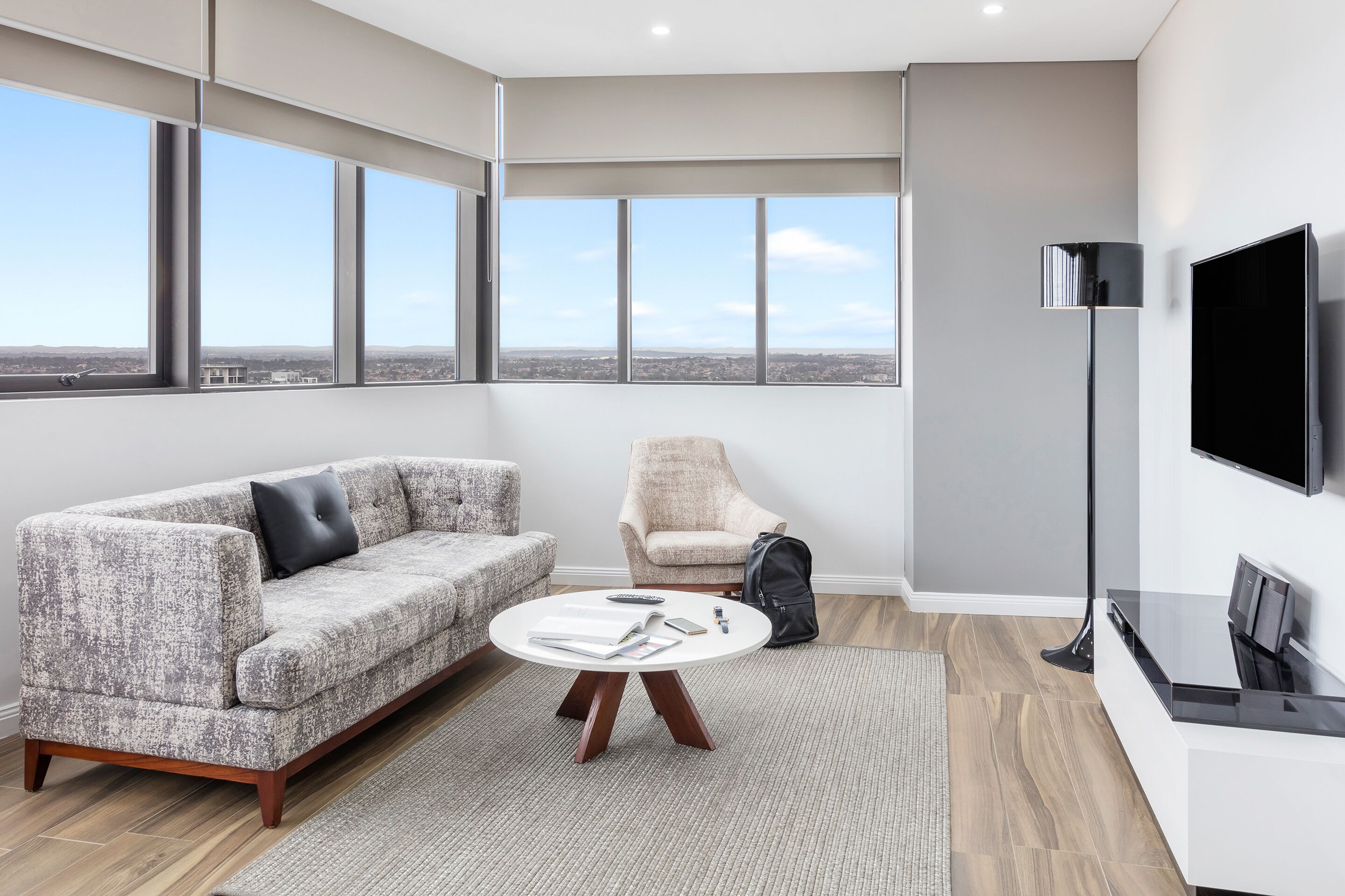 Una sala de estar moderna con un sofá gris, una mesa de centro redonda, un sillón beige, una lámpara de pie negra y un televisor de pantalla plana.