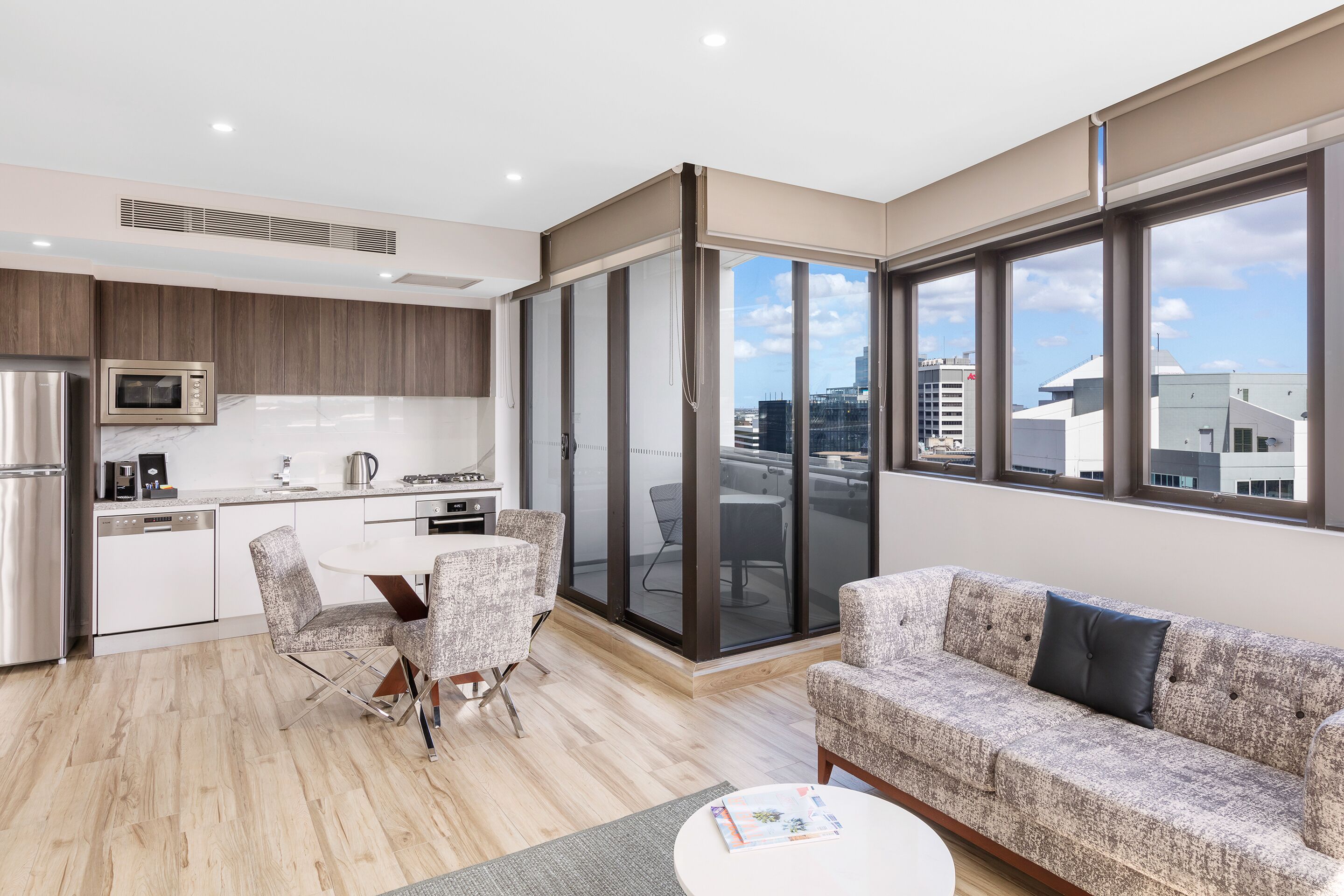 A modern apartment with a kitchen, dining area, and living room. It features a large sliding glass door to a balcony, a sofa, and a coffee table.