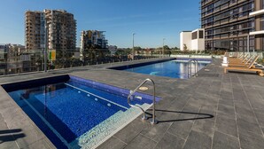 Un área de piscina en la azotea con dos piscinas, sillas de descanso y vistas de edificios circundantes.