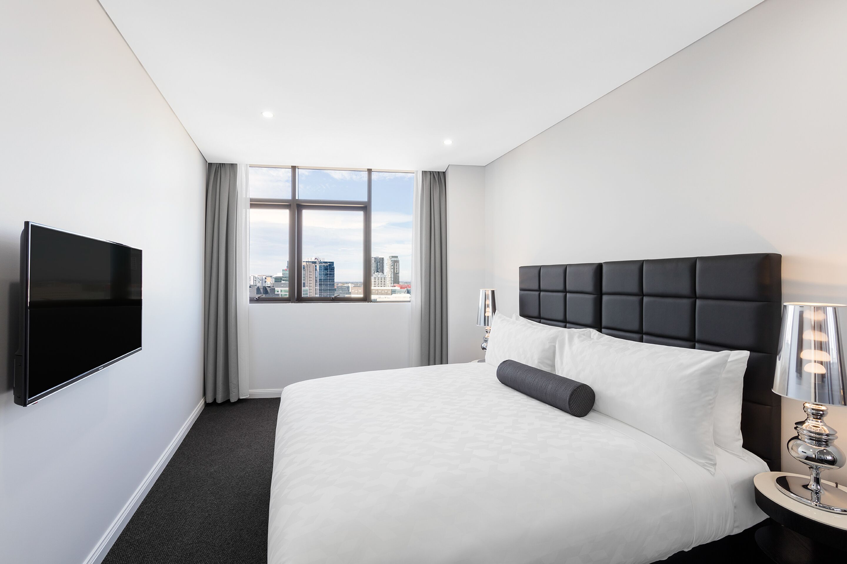 Habitación de hotel con una cama grande, un televisor de pantalla plana, una ventana con vista a la ciudad y una mesita de noche con una lámpara.
