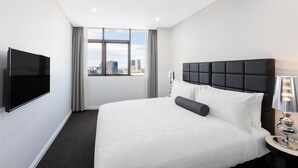 Habitación de hotel con una cama grande, un televisor de pantalla plana, una ventana con vista a la ciudad y una mesita de noche con una lámpara.